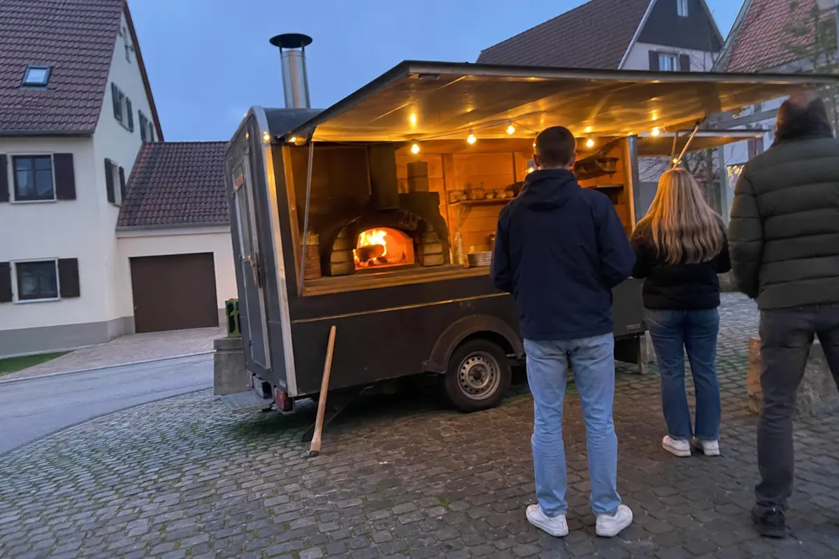 Das Pizzamobil am frühen Abend auf dem Dorfplatz, Holzofen glüht, ein paar Gäste warten an der Klappe